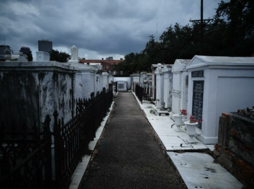 A narrow path between white tombs in a cemetery, under a cloudy sky, with iron fences lining the sides.