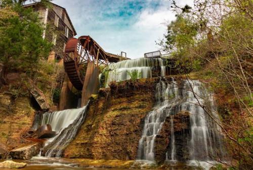 A scenic view of a watermill with a large wheel, cascading waterfalls, and lush greenery surrounding the area.