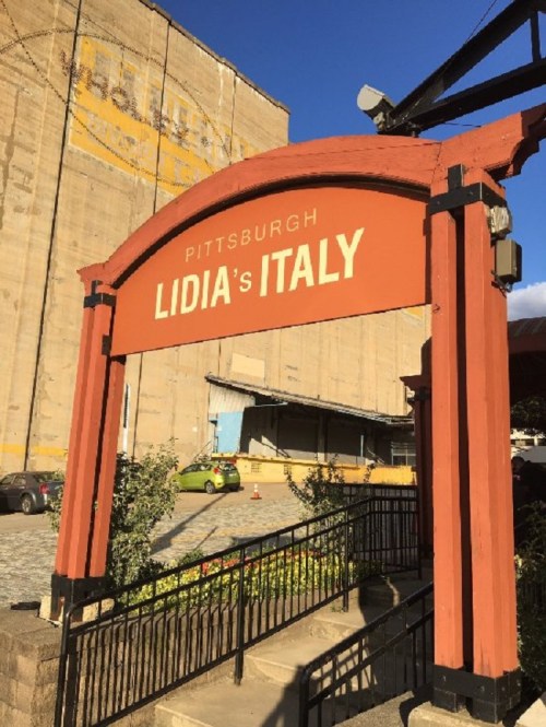 Sign for "Lidia's Italy" in Pittsburgh, featuring a decorative archway and surrounding greenery.