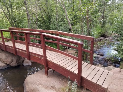 A wooden bridge spans a small stream, surrounded by lush greenery and rocky terrain.