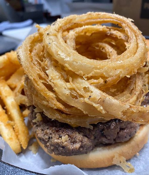 A close-up of a burger topped with crispy onion rings, served with a side of fries on a plate.