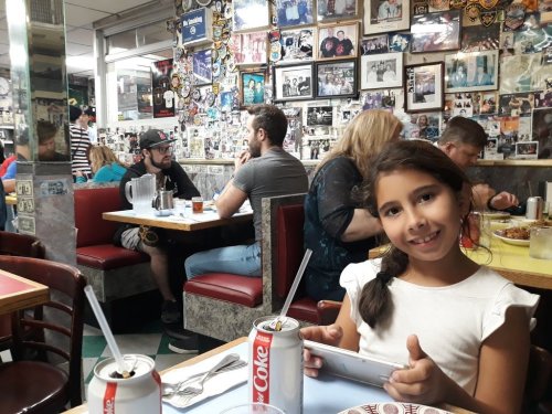 A young girl smiles while holding a device in a busy diner filled with people and photos on the walls.