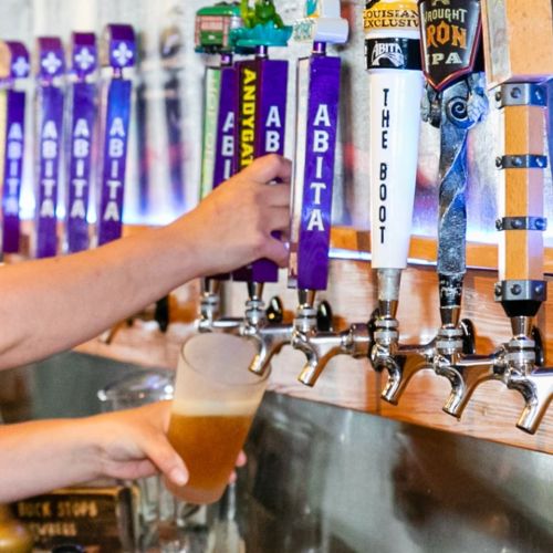A hand reaches for a beer tap, with various colorful tap handles displayed above a glass being filled with beer.