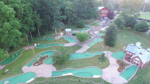 Aerial view of a mini-golf course with winding paths, greenery, and a rustic building in the background.