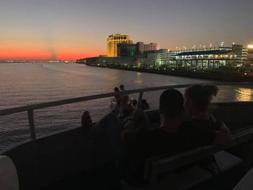 A couple relaxes on a boat, enjoying a sunset over the water with city lights in the background.