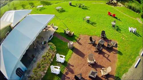 Aerial view of a grassy outdoor area with seating, tables, and a tent, surrounded by trees and a peaceful landscape.