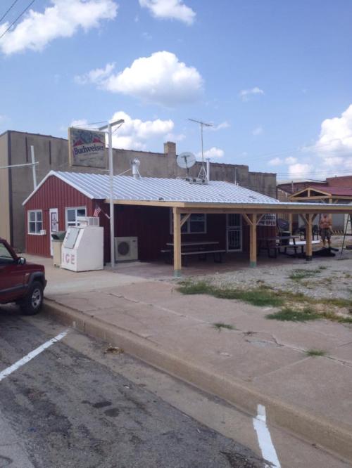 A small red building with a metal roof, outdoor seating, and a sign for Budweiser, set in a rural area.