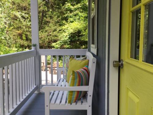 A cozy porch with a white bench and colorful pillows, surrounded by greenery and a bright yellow door.