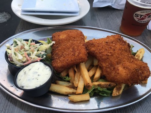 A platter with two crispy fish fillets on a bed of fries, accompanied by coleslaw and dipping sauce.