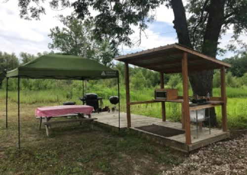 A picnic area with a green canopy, picnic table, and a cooking station under a wooden shelter in a grassy field.