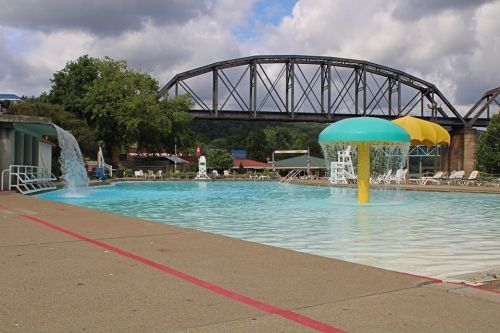 A swimming pool with a water feature, colorful umbrellas, and a bridge in the background under a cloudy sky.