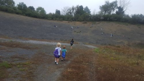 Two children walk on a gravel path towards a sloped, rocky hill with trees in the background on a cloudy day.