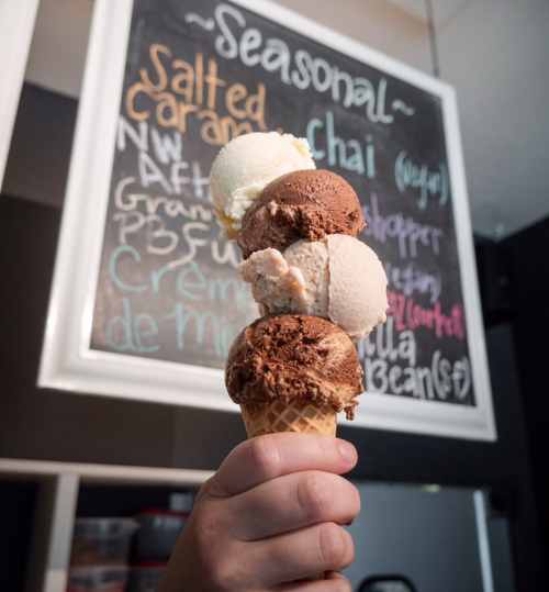A hand holding a cone with four scoops of ice cream in various flavors, with a chalkboard menu in the background.