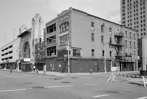 Black and white image of an old building with a theater entrance, fire escape, and nearby parking structure.