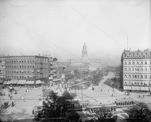 Historic black-and-white photo of a busy city square with buildings, a monument, and people walking around.