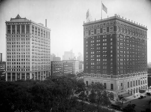 Historic black-and-white photo of two tall buildings, one with an American flag, surrounded by trees and cityscape.
