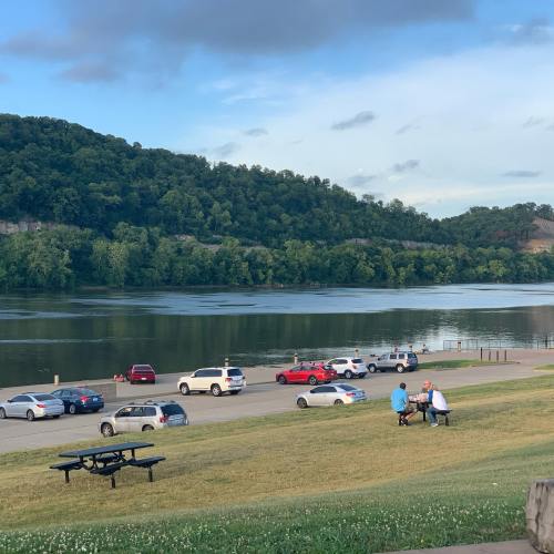A serene river scene with cars parked along the shore, people sitting at a picnic table, and lush green hills in the background.