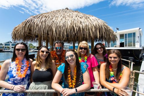 A group of eight people in tropical attire, smiling and posing under a thatched roof by the water on a sunny day.