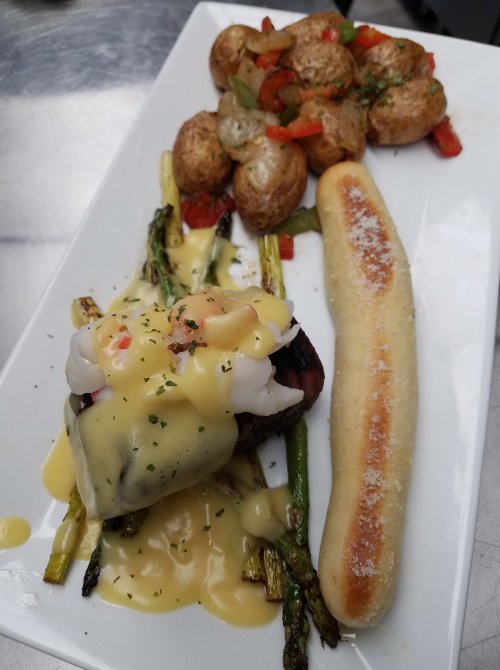 A plated meal featuring steak topped with shrimp and hollandaise, asparagus, roasted potatoes, and a breadstick.