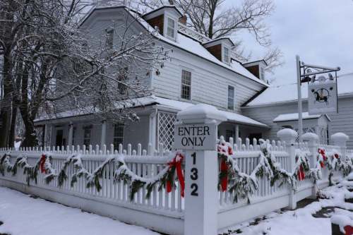 A white house with snow-covered roof and festive decorations, surrounded by a white fence and winter scenery.