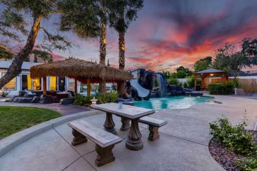 A backyard oasis featuring a pool, slide, thatched gazebo, and vibrant sunset sky.