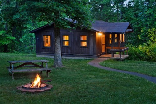 A cozy cabin surrounded by trees, with a fire pit in front and warm light glowing from the windows at dusk.