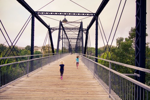 Two children run on a wooden bridge surrounded by trees, with a cloudy sky above.