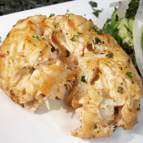 A close-up of a golden-brown crab cake, garnished with parsley, served alongside broccoli on a white plate.