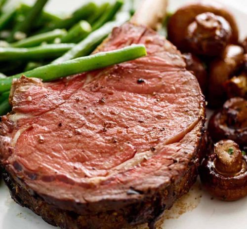 A juicy ribeye steak with green beans and sautéed mushrooms on a white plate.