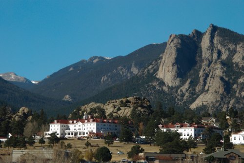 A large white hotel with a red roof nestled among trees, set against a backdrop of rocky mountains and clear blue sky.