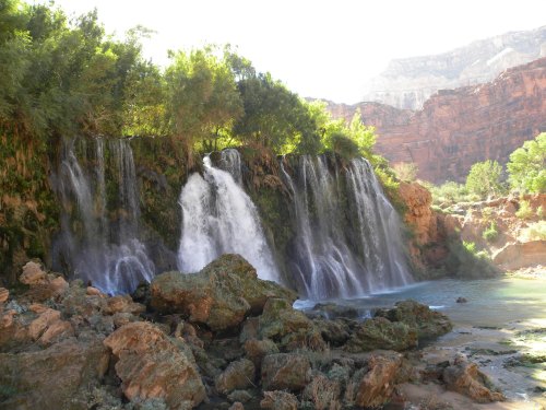 A serene waterfall cascades over rocks, surrounded by lush greenery and red rock formations in a sunny canyon.