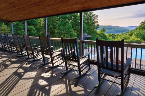 A row of rocking chairs on a porch overlooking a scenic view of green hills and a pool in the distance.