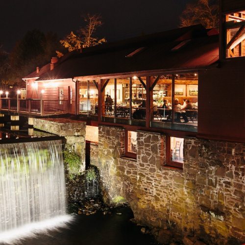 A cozy restaurant by a waterfall at night, with warm lights illuminating the interior and guests dining inside.