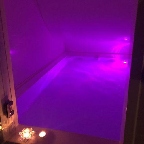A serene, dimly lit room featuring a purple-lit soaking tub and candles, creating a relaxing atmosphere.