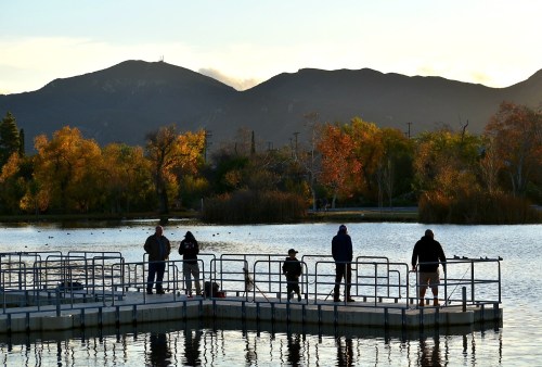 People fishing on a dock at sunset, surrounded by autumn trees and mountains in the background.