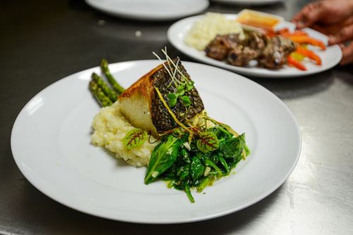 A beautifully plated dish featuring seared fish on risotto, garnished with greens and asparagus, with another dish in the background.