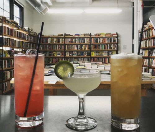 Three colorful cocktails on a table in a cozy bookstore, with shelves of books in the background.