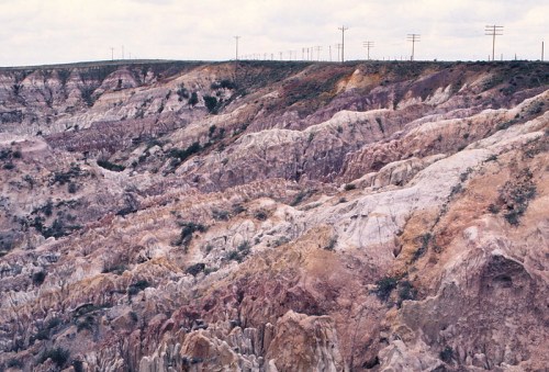 Colorful, rugged landscape of eroded hills with sparse vegetation and distant power lines under a cloudy sky.