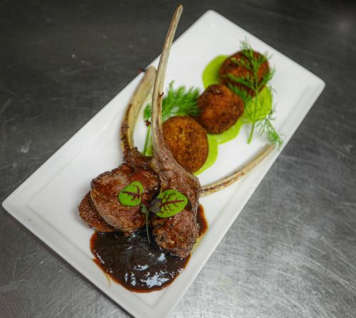 A gourmet dish featuring lamb chops, green sauce, and croquettes, garnished with herbs on a white plate.