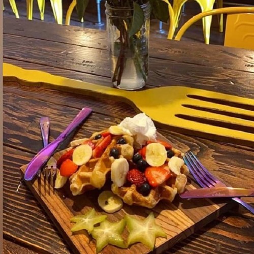 A wooden plate with a waffle topped with fruits, whipped cream, and starfruit, set on a rustic table with colorful decor.