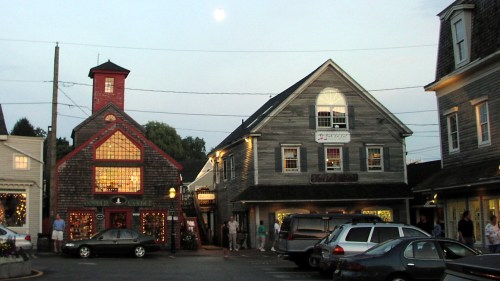 A quaint town square at dusk, featuring charming shops and a full moon in the sky. Cars are parked nearby.