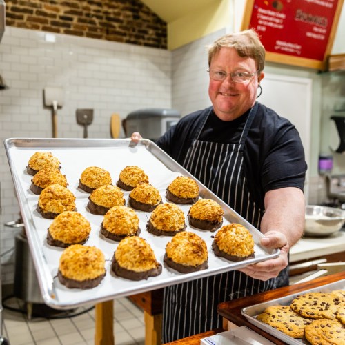 A smiling baker holds a tray of coconut macaroons dipped in chocolate, with more cookies in the background.