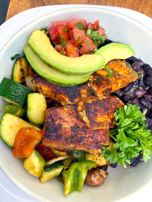 A colorful bowl featuring seasoned fish, black beans, fresh tomatoes, avocado slices, and assorted grilled vegetables.