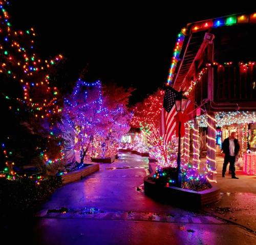 Colorful holiday lights illuminate a pathway and trees, with a building decorated and an American flag displayed.