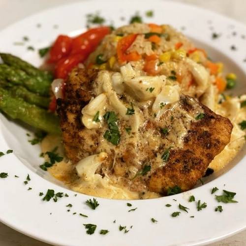 A plate of seasoned fish topped with creamy sauce, served with rice, mixed vegetables, and asparagus, garnished with parsley.
