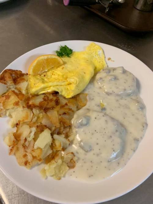 A plate of breakfast featuring an omelet, hash browns, and biscuits smothered in creamy gravy, garnished with an orange slice.
