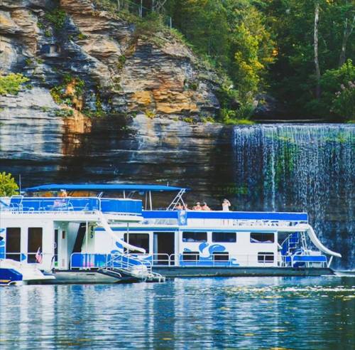 A houseboat anchored near a rocky shore with a waterfall cascading in the background, surrounded by lush greenery.
