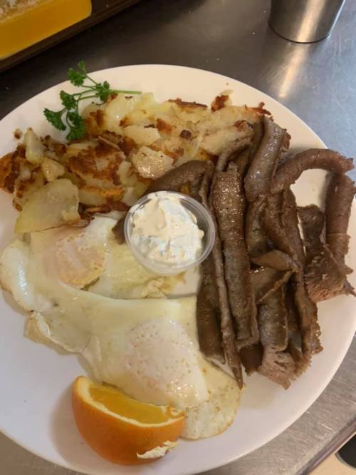A plate of breakfast featuring eggs, seasoned beef strips, hash browns, and a slice of orange with a small cup of sauce.