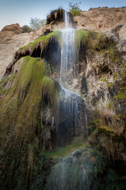 A cascading waterfall flows down a rocky cliff, surrounded by lush green moss and vegetation.