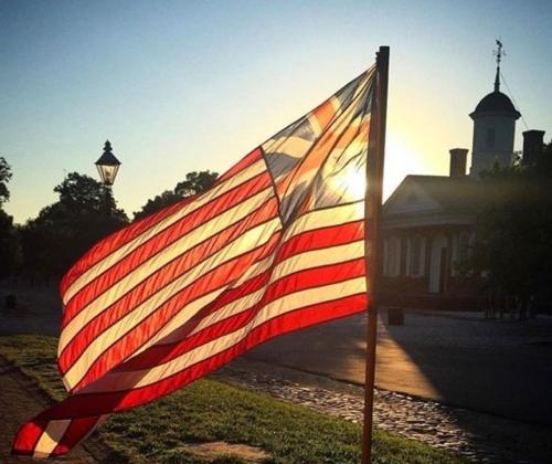 A U.S. flag waves in the foreground with a sunset and historic building in the background.